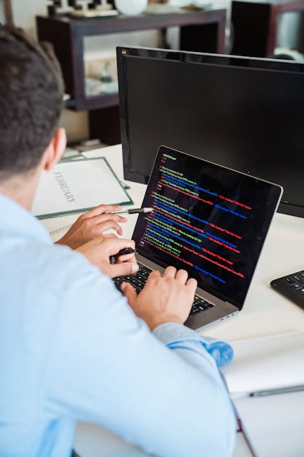 A person wearing a light blue shirt is seated at a white desk, working on a laptop that displays lines of colorful programming code on its screen. The individual is holding a stylus in one hand and us