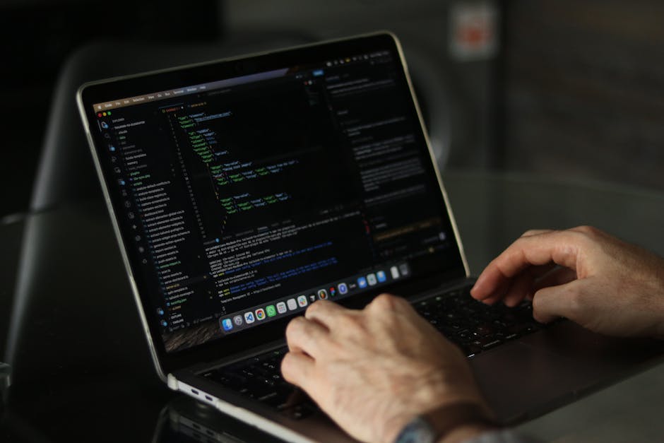 A person wearing a light blue shirt is seated at a white desk, working on a laptop that displays lines of colorful programming code on its screen. The individual is holding a stylus in one hand and us