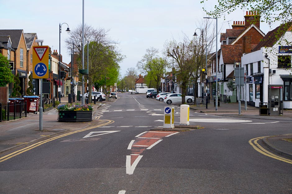 A quiet residential street in Woking with small shops and houses on either side, featuring a give way sign, street lamps, and trees with some greenery. The road has markings indicating a roundabout ah