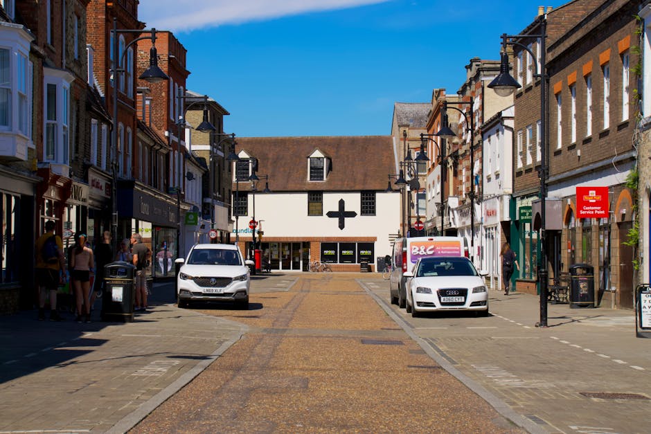 A quiet residential street in Woking with small shops and houses on either side, featuring a give way sign, street lamps, and trees with some greenery. The road has markings indicating a roundabout ah